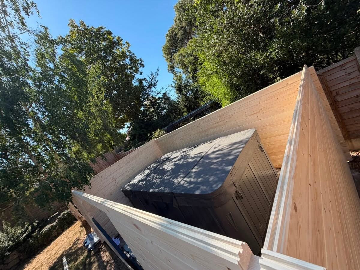A dark gray hot tub or spa cabinet placed outdoors on a light wood deck, surrounded by trees and a blue sky.