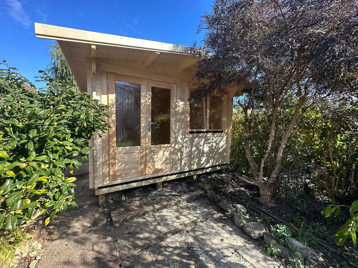 A small wooden cabin or shed with a light natural wood exterior, large glass doors, and windows, situated outdoors on a stone path surrounded by green shrubs and trees under a blue sky.