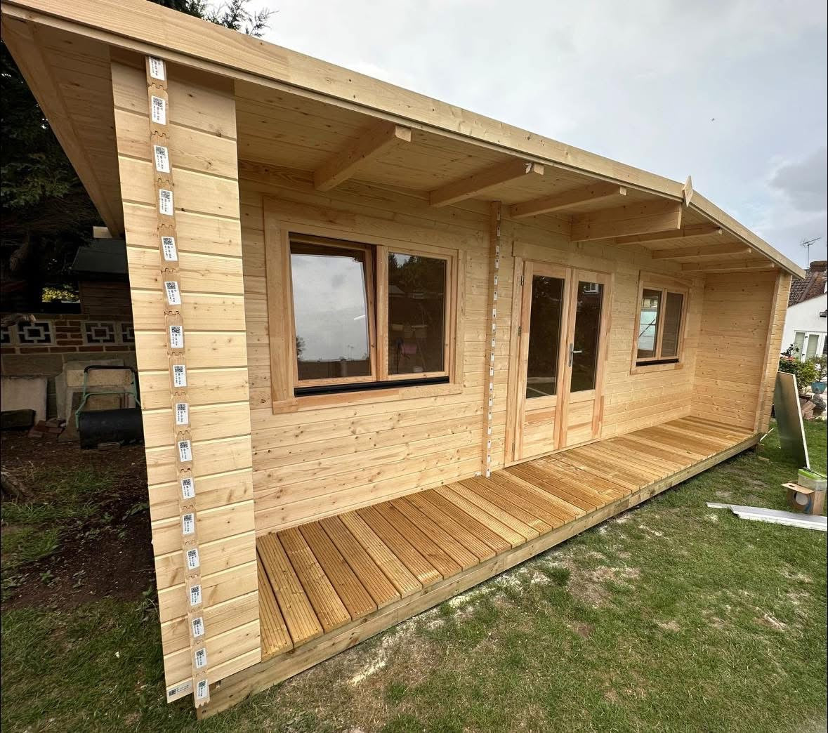 A light natural wood log cabin with a matching wooden deck and porch, featuring double glass doors and windows, set outdoors on a grassy lawn under an overcast sky.