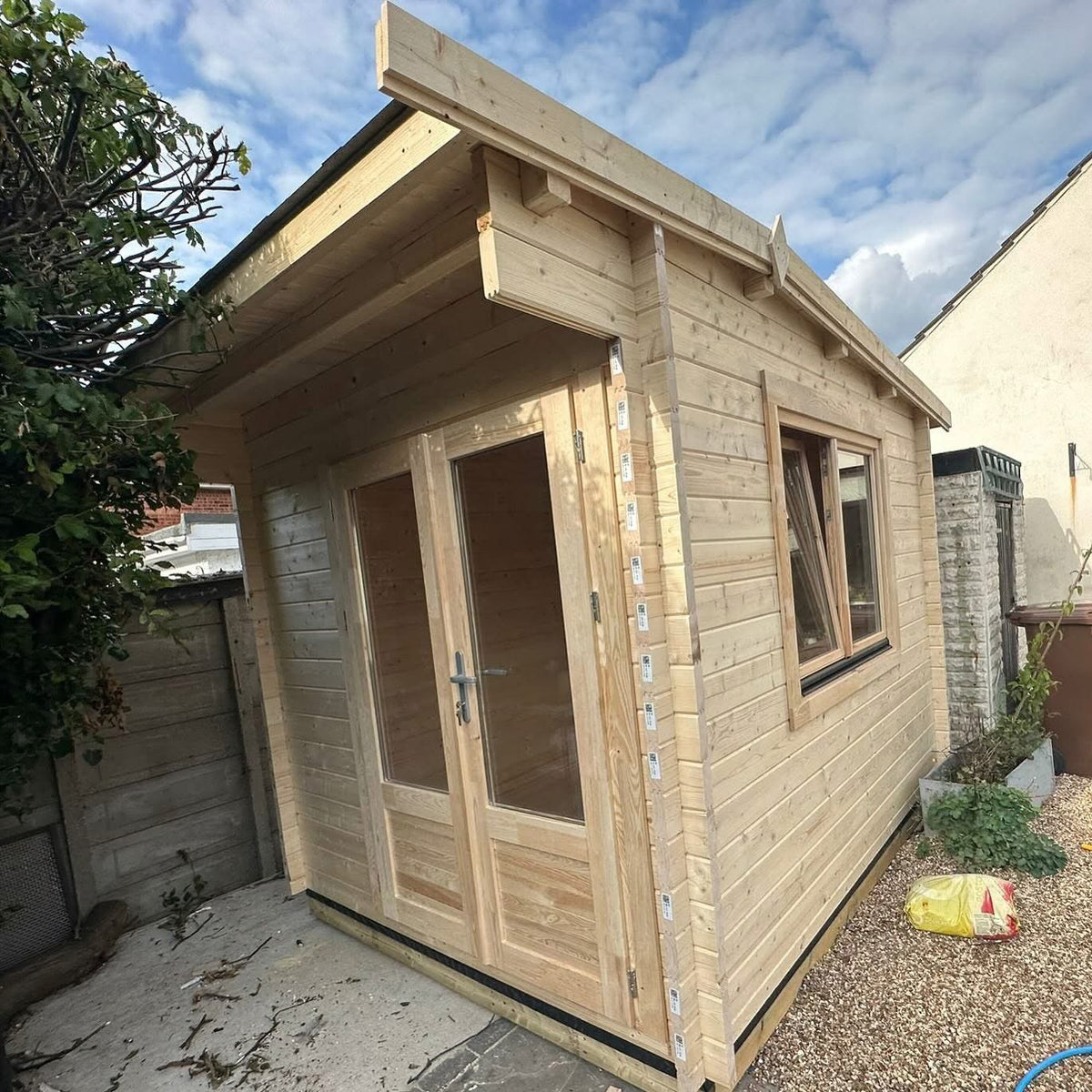 A small wooden garden shed with light natural wood paneling, double glass doors, and a single window, photographed outdoors on a gravel and concrete patio with trees and neighboring buildings visible.