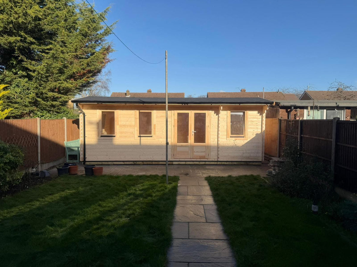 A single-story wooden garden shed with light beige wood paneling and brown double doors, situated in a backyard with a stone pathway, green lawn, and wooden fences under a clear blue sky.