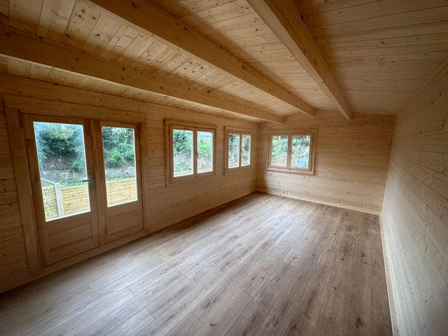 An empty rustic wooden room with light natural wood plank walls, ceiling, and floor, featuring several tall windows looking out onto greenery.