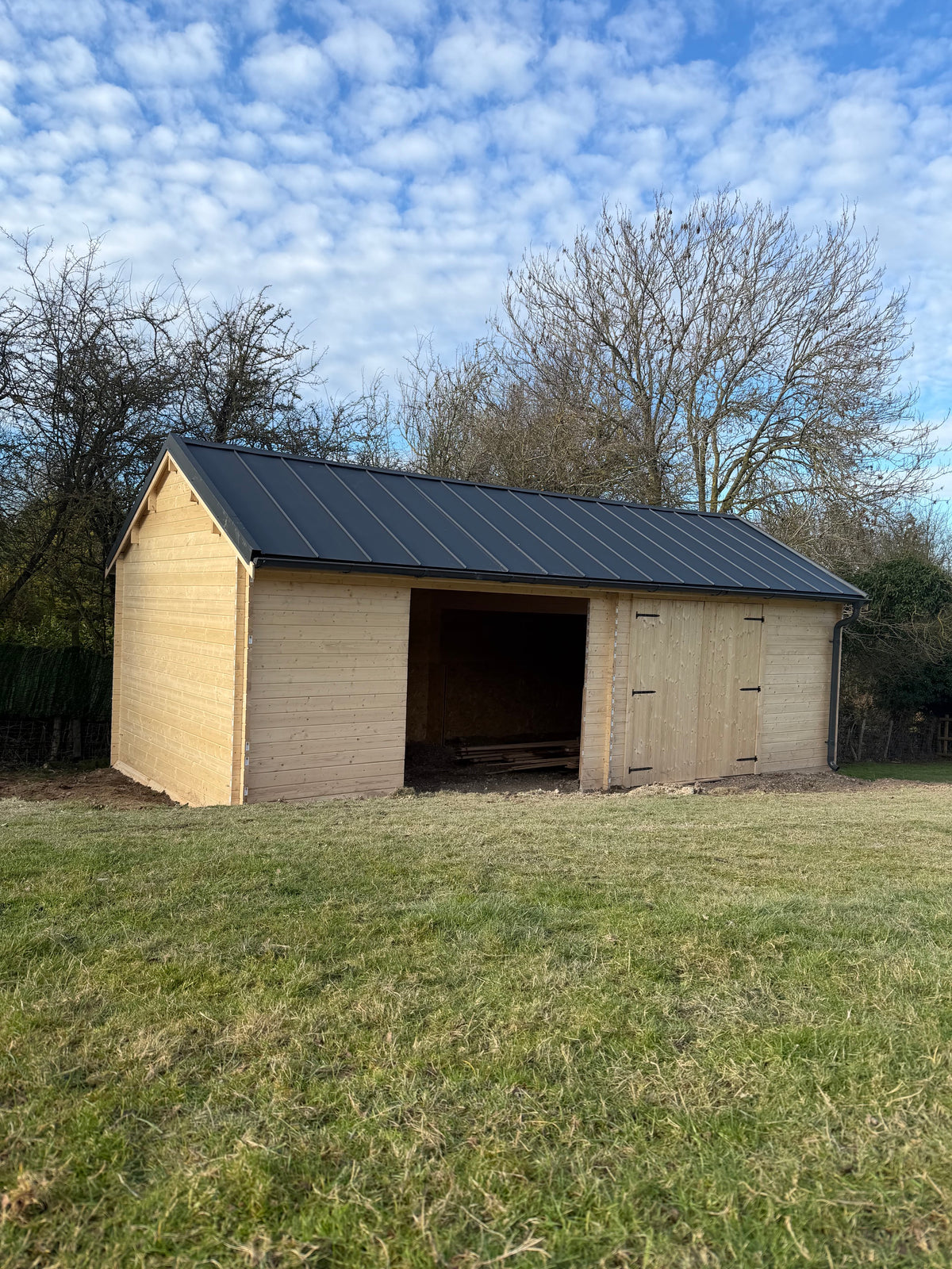 A small wooden shed or stable with a dark metal roof and open front, photographed outdoors on a grassy field with trees and a blue cloudy sky in the background.