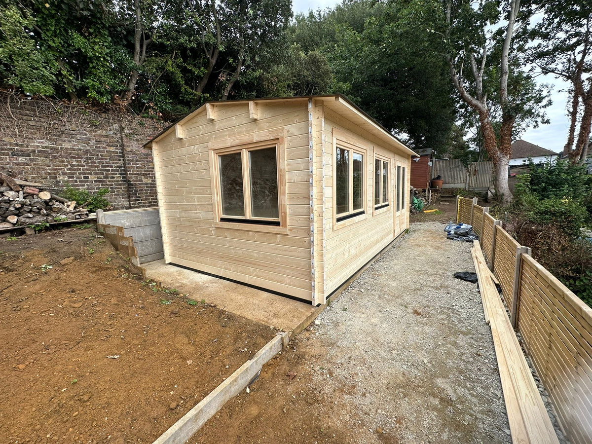 A small wooden garden shed with light natural wood paneling and multiple windows, shown outdoors on a gravel path next to a dirt area and a wooden fence.