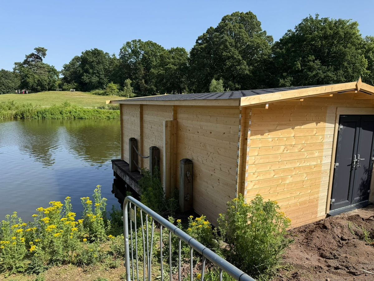 A light natural wood floating home or cabin with a dark metal roof and double doors, situated on a body of water with trees and grass in the background.