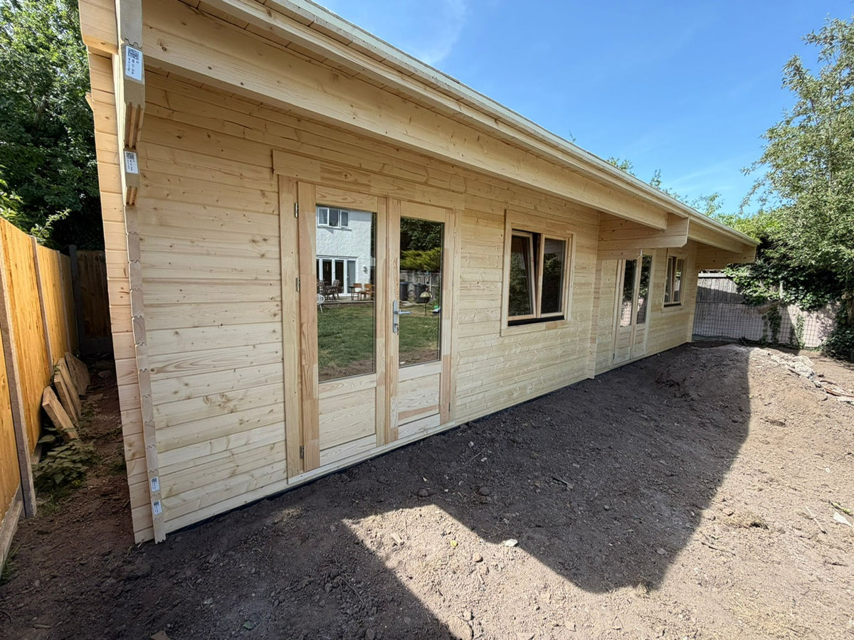A light natural wood log cabin or extension with glass double doors and windows, shown outdoors on a dirt ground with trees and a fence in the background.