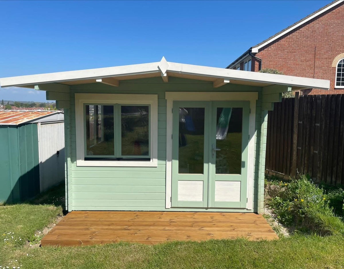 A small light green garden shed with white trim, double glass doors, a single window, and a wooden deck in a backyard.