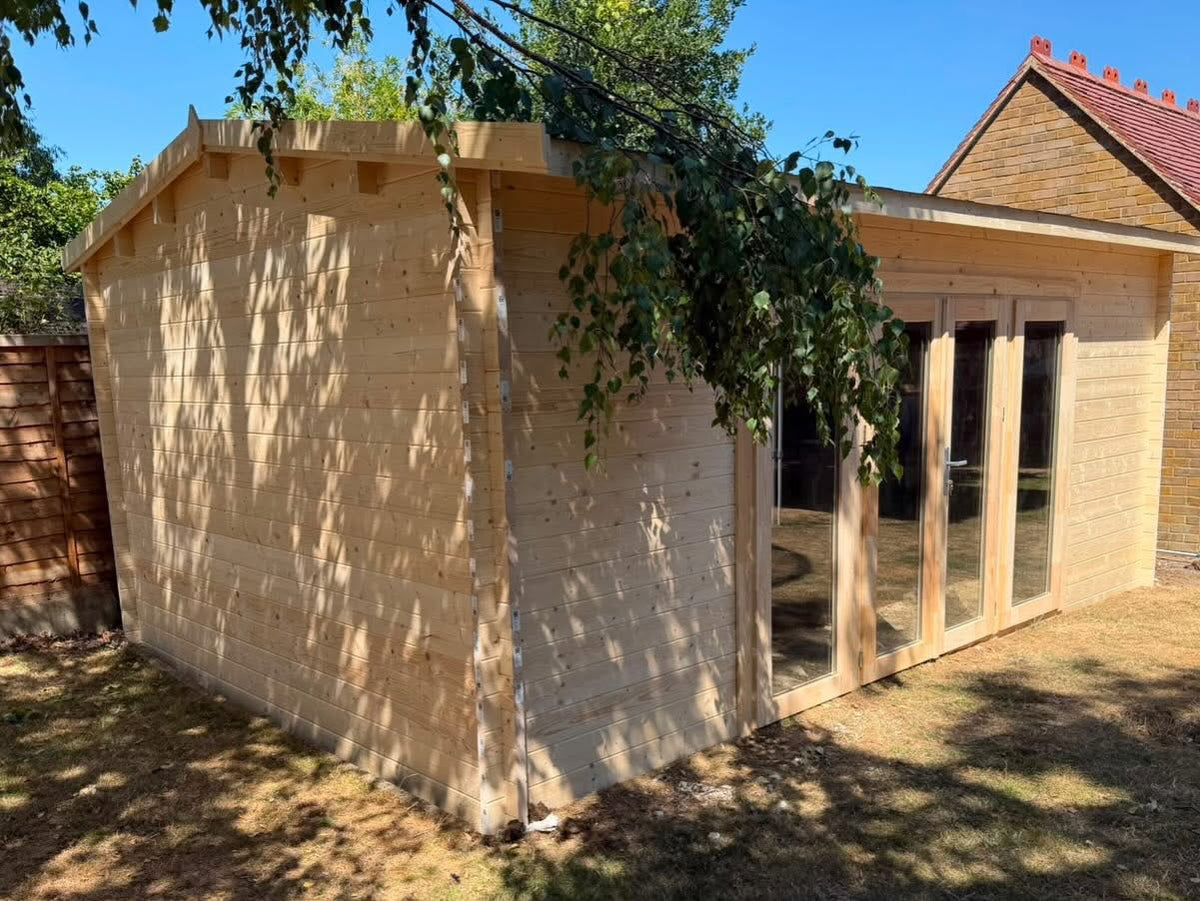 A light natural wood garden shed with a pitched roof and glass door/window panels, shown outdoors on grass with tree shadows and a neighboring house in the background.