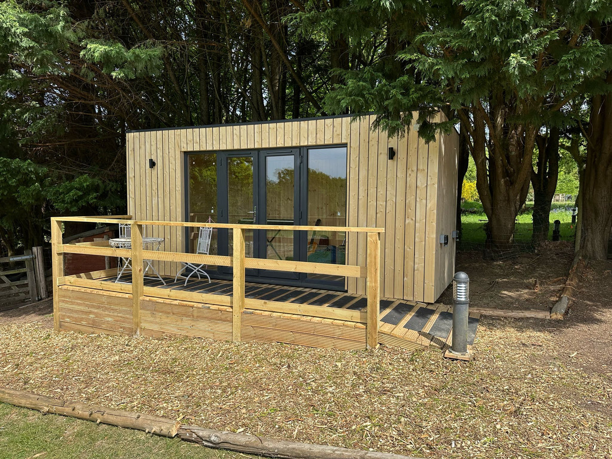 A modern wooden cabin with a raised wooden deck and railing, featuring dark-framed glass doors, set in a wooded outdoor area with trees and mulch.