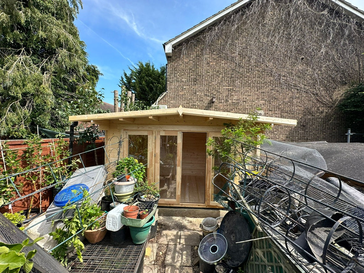 A small wooden garden shed with glass doors and windows, situated in a backyard with plants, pots, a brick wall, and a house in the background.